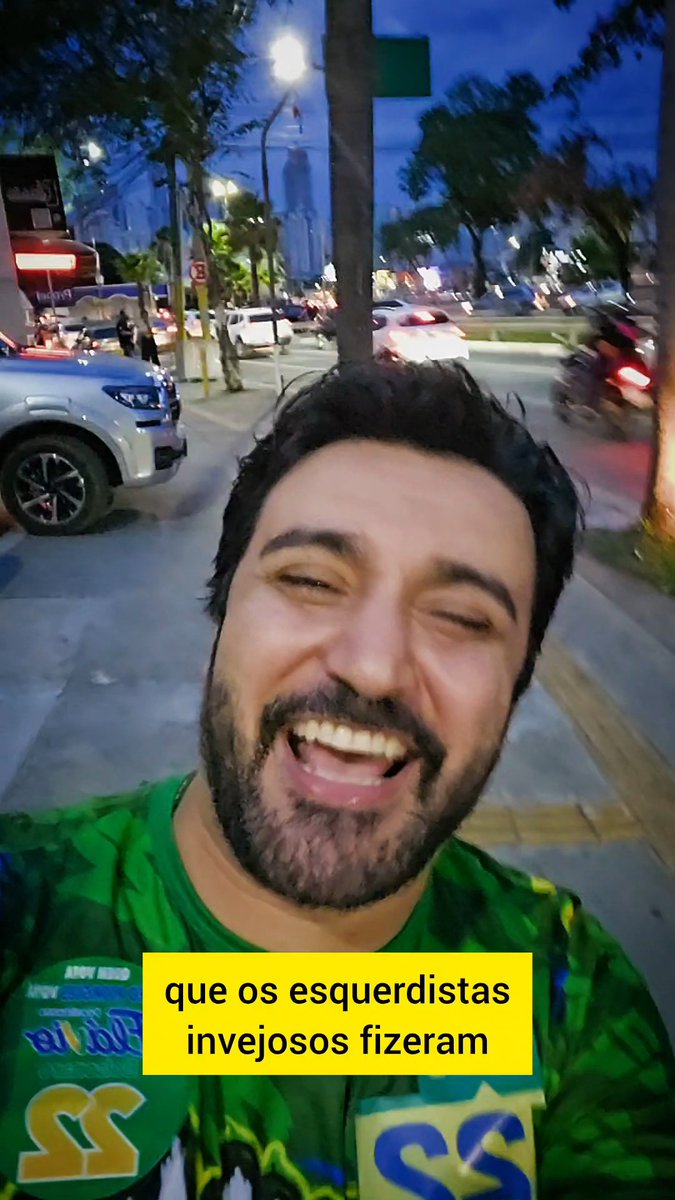 Vertical video still of a smiling man on a city sidewalk at dusk, wearing a green shirt, with a yellow Portuguese subtitle box reading “que os esquerdistas invejosos fizeram.”