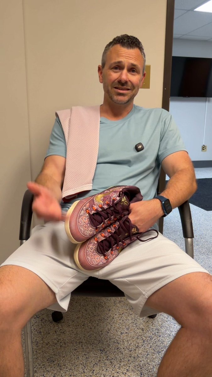 A seated man in a light blue shirt and white shorts holds up a pair of maroon patterned soccer cleats, with a pink towel draped over his shoulder and a small microphone clipped to his shirt.