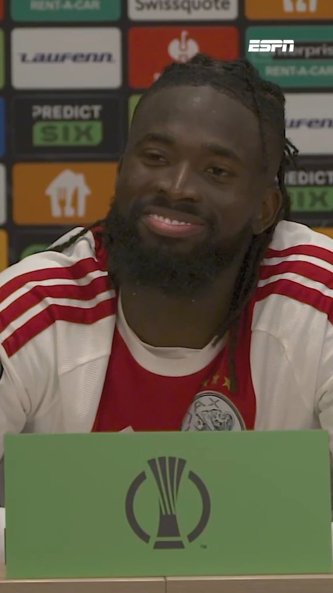 A smiling Black man in a red-and-white football jersey sits behind a green microphone board with the DFB-Pokal trophy logo. An ESPN interview backdrop shows “Swissquote,” “PREDICT SIX,” and other sponsor text, fitting the post’s comment about Tolu being in strong form away from the field.