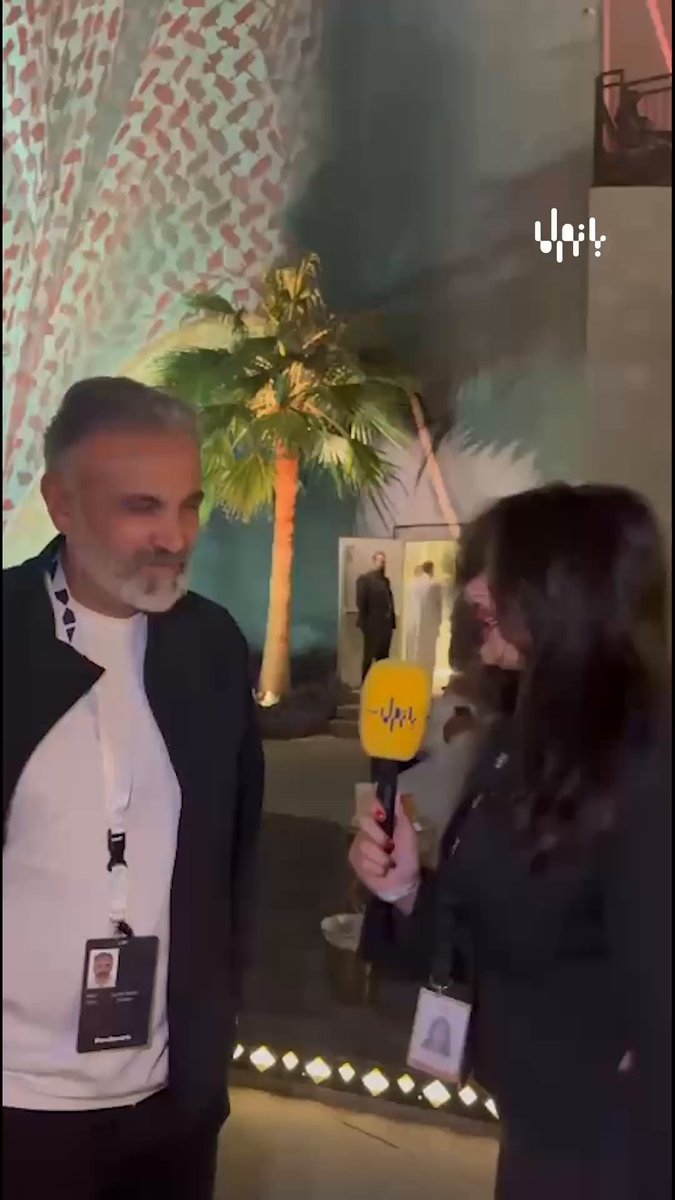 A woman holding a yellow microphone interviews a gray-haired man wearing an event badge in front of a palm tree and a patterned wall; a white Arabic logo appears at the upper right.