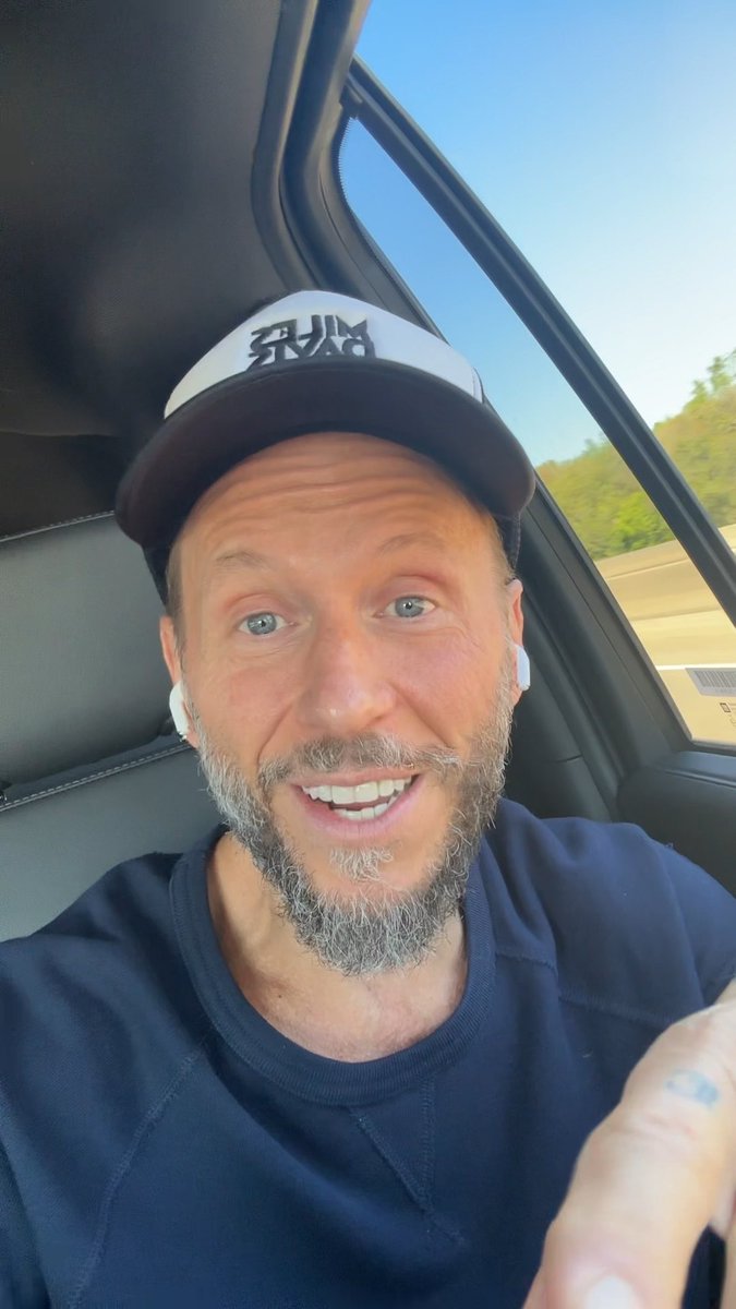 A smiling bearded man wearing a white earbud, navy shirt, and black-and-white cap films himself from the driver’s seat of a car.