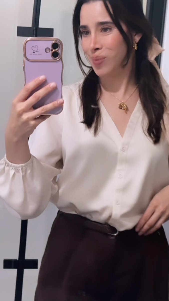 A woman takes a mirror selfie while wearing a white blouse, dark high-waisted bottoms, gold jewelry, and a light bow in her dark hair.