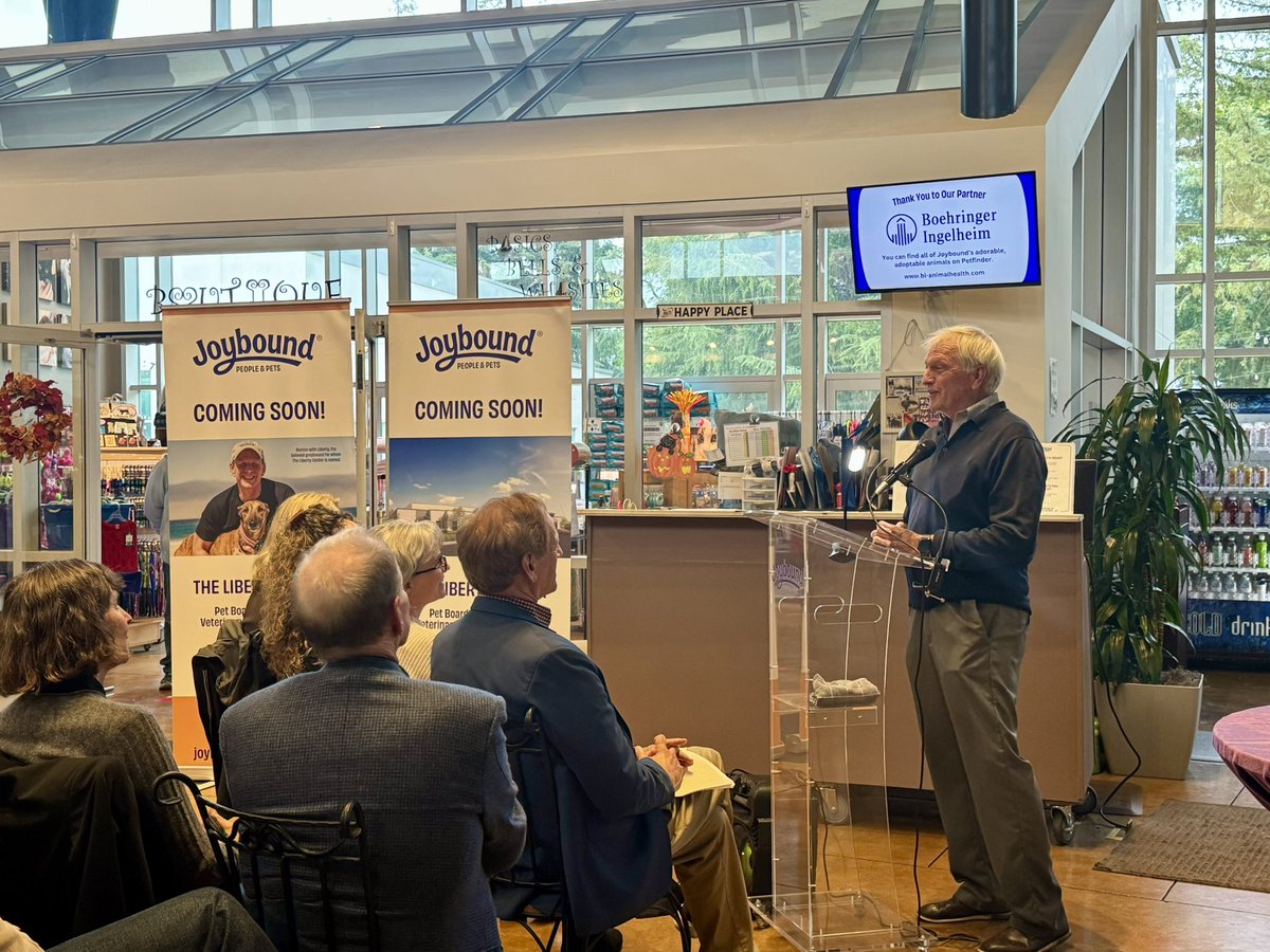 An older man speaks at a clear lectern to a seated audience inside a bright retail space. Two promotional banners stand behind the audience, and a wall-mounted screen displays a sponsor message. There is text throughout the scene.