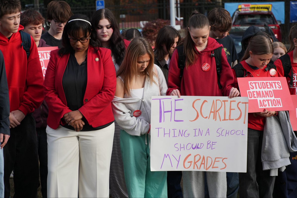 A group of students and an adult stand outdoors at a demonstration, looking down and holding signs. Many wear red clothing and button badges. The signs contain handwritten and printed text.