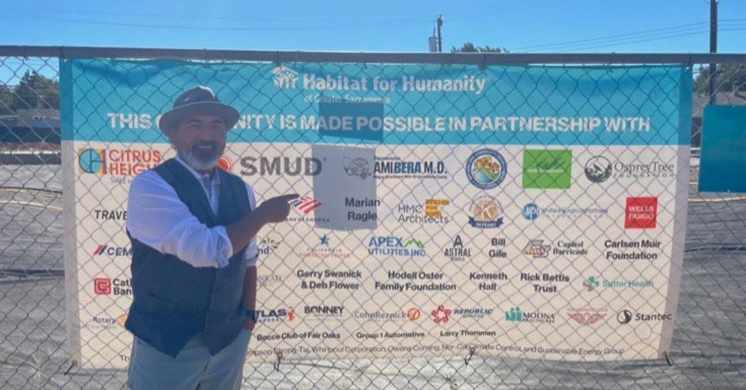 A man in a hat and vest stands beside a chain-link fence, pointing toward a large Habitat for Humanity banner covered with sponsor logos and text.