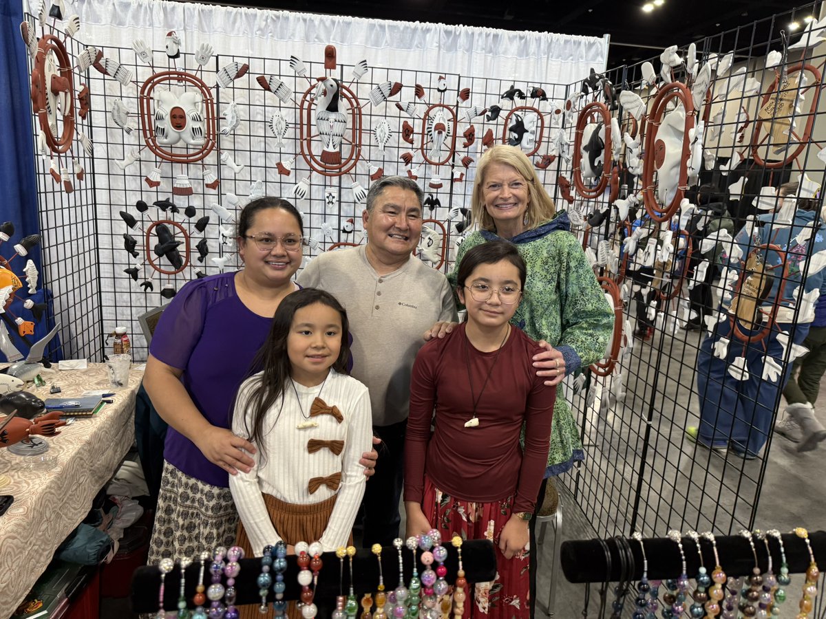 A group of five people pose together at an indoor craft or art market booth. Behind them, a grid display is covered with handmade masks and sculptural art, while tables and stands in front hold jewelry and other crafts. There is some text visible on clothing and display items.