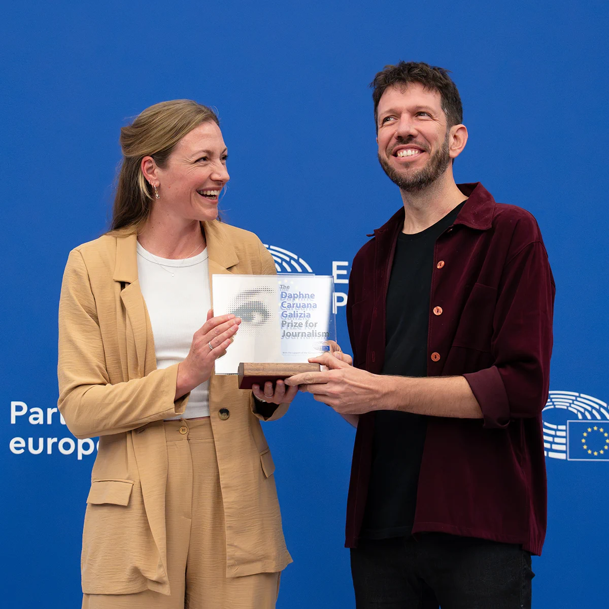 Journalists Giacomo Zandonini and Maite Vermeulen jointly holding the Daphne Caruana Galizia Prize for Journalism trophy in front of a blue European Parliament backdrop.