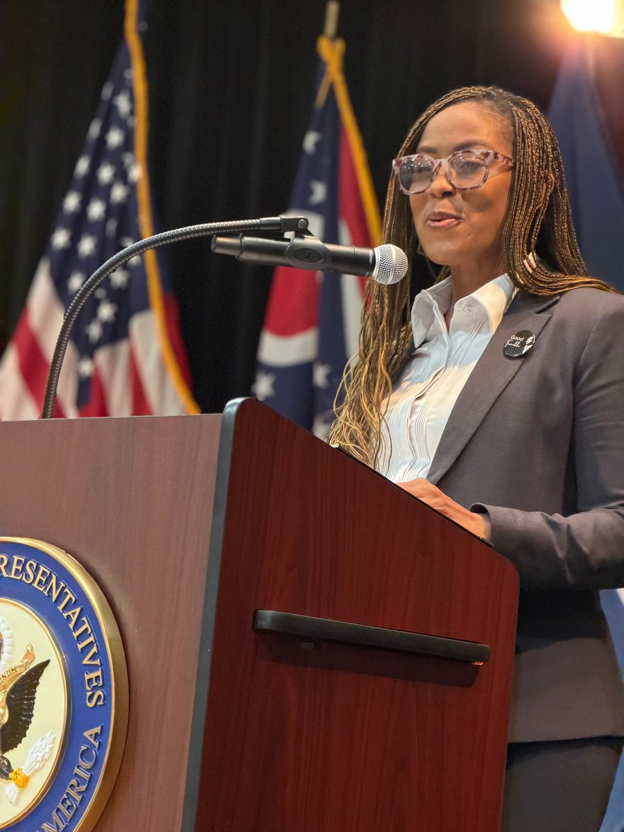 A woman wearing glasses and a dark suit speaks at a wooden podium with a microphone. Several flags and a government-style seal are visible behind and on the podium. There is text on a pin and seal.