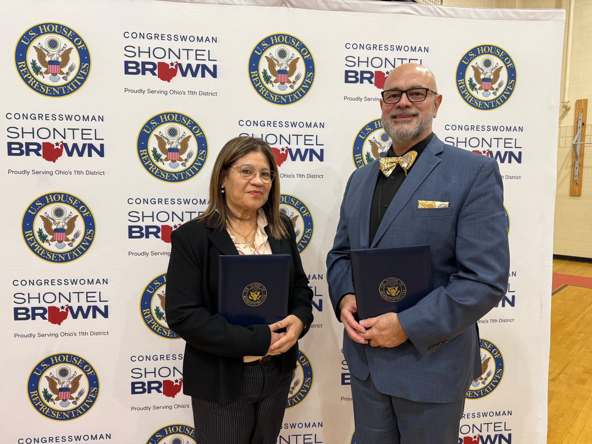 A woman and a man stand together for a posed photo in front of a repeating congressional campaign backdrop. Each holds a dark blue folder with a gold seal. Text and official-looking emblems appear across the backdrop.