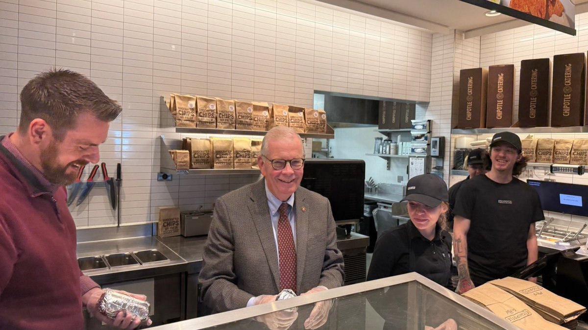 Several people stand inside a fast-casual restaurant behind a glass food counter. A man on the left holds a foil-wrapped item, an older man in a suit stands in the center, and two employees are on the right. Shelves of packaged chips and restaurant equipment are visible in the background. There is text on the packages and containers.