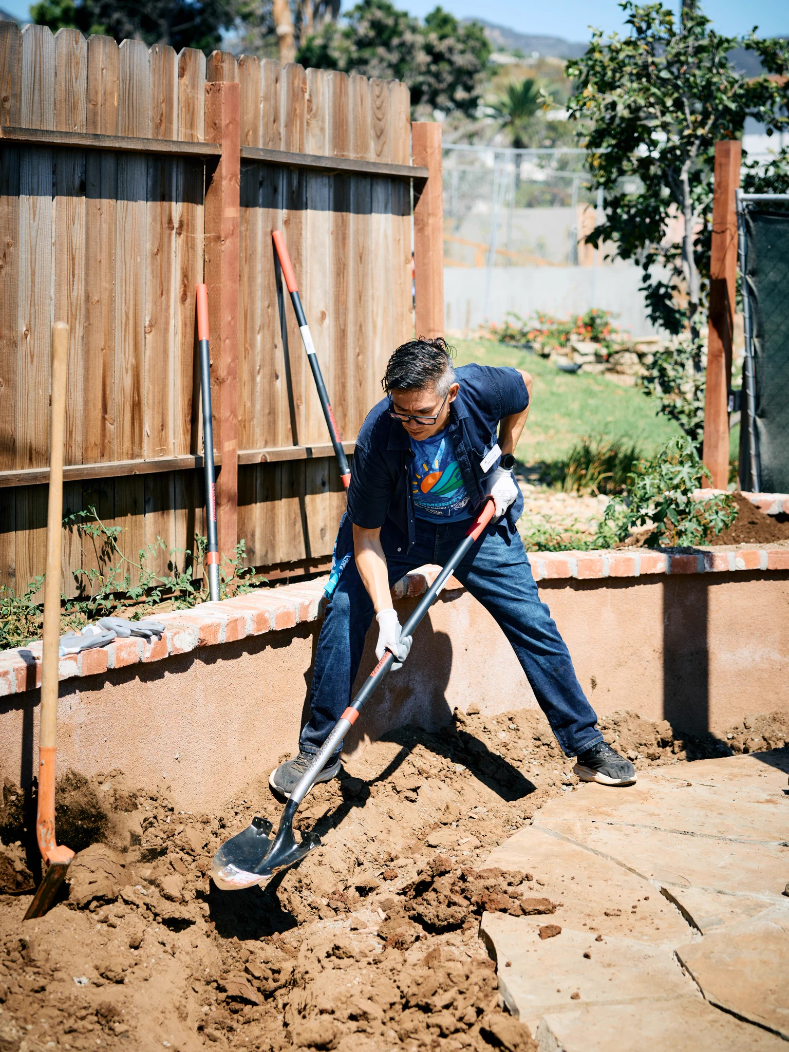 A person is gardening, using a shovel to dig in a soil area near a wooden fence. The garden has a brick border and is next to a paved path. There are trees and plants in the background.