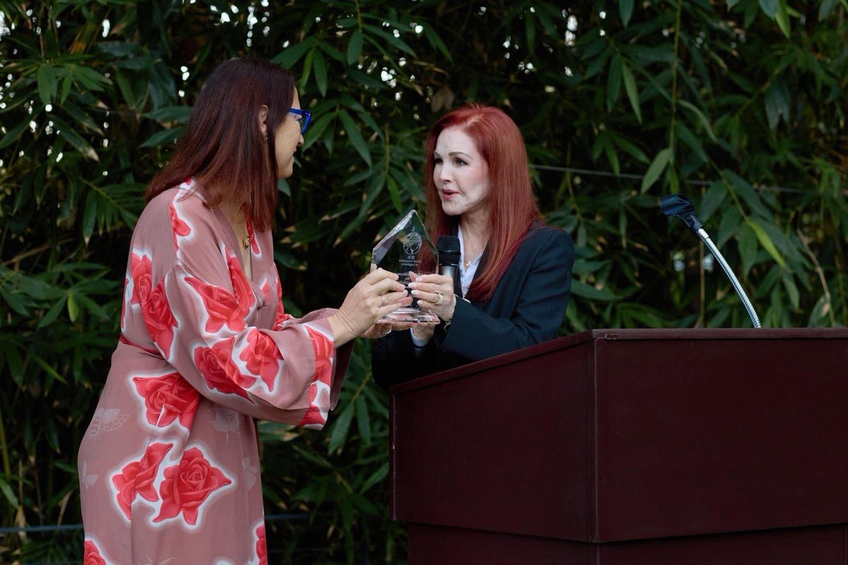 Two women stand at an outdoor event as one presents the other with a clear award. The recipient holds a microphone beside a wooden podium, with dense green foliage in the background. The award has some text.