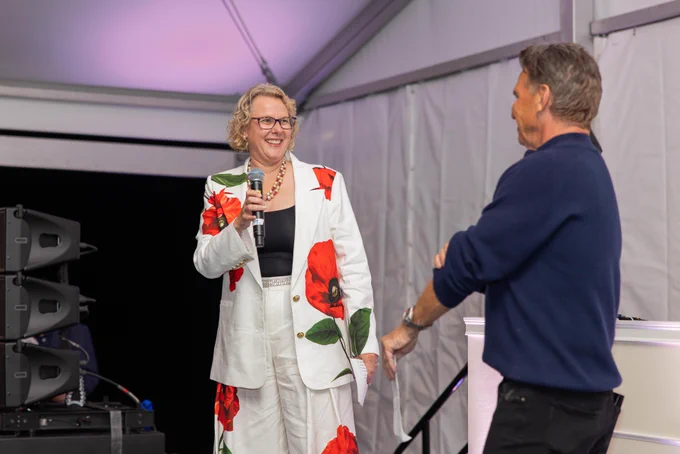 A smiling, middle-aged woman with blonde hair and glasses wears a white suit with red floral patterns. She holds a microphone, speaking to a man in a dark blue sweater, seen from behind, on a stage inside a tent.