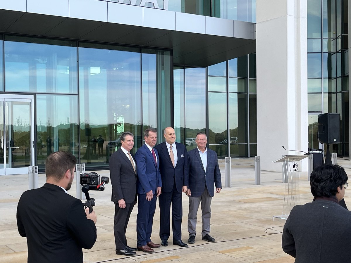 Four men in business attire pose for a photograph outside a modern glass-fronted building while a photographer aims a camera at them. A podium and speaker are nearby. No prominent text is visible.