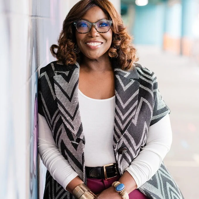 Kinitra Brooks smiles at the camera while leaning against a wall. She wears glasses, a white top, and a black-and-white patterned vest.