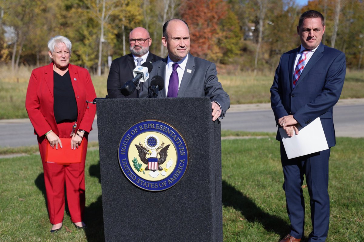 Four people stand outdoors at a press event. A man in a suit speaks at a podium with microphones and an official seal, while the others stand nearby. There is text on the podium seal.