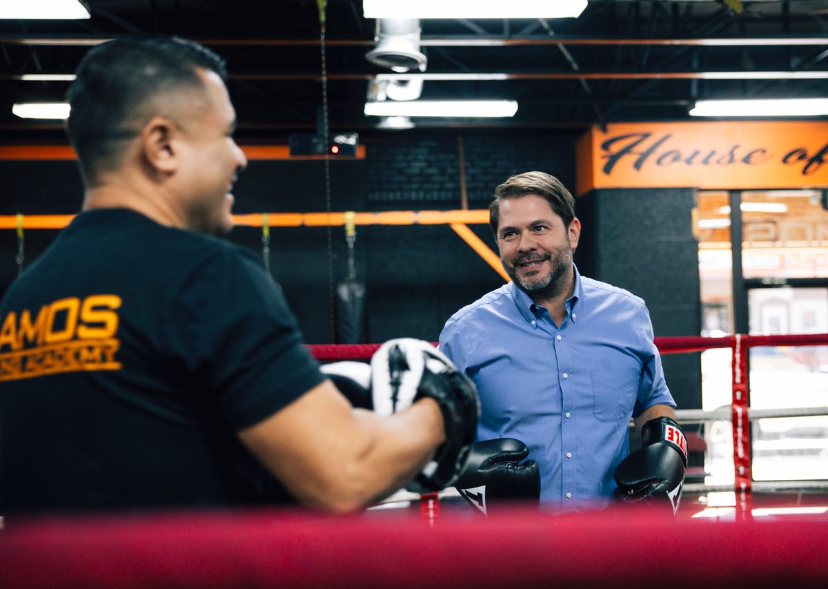 Two men are talking inside a boxing gym. One wears boxing gloves and a blue shirt, while the other, seen from behind, wears a dark T-shirt and holds training mitts. They stand in a boxing ring. There is text on the shirt and a sign in the background.