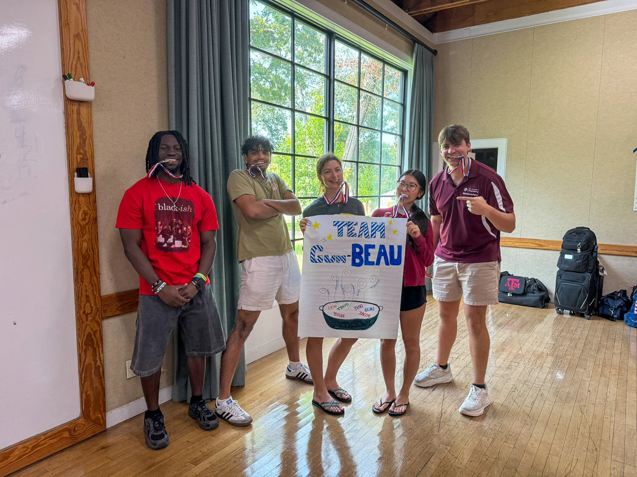 A group of five people stands indoors on a wooden floor, near a tall window. They are holding a colorful sign that reads "TEAM BEAU." Four individuals are wearing medals around their necks. Behind them, a backpack is visible on the floor.
