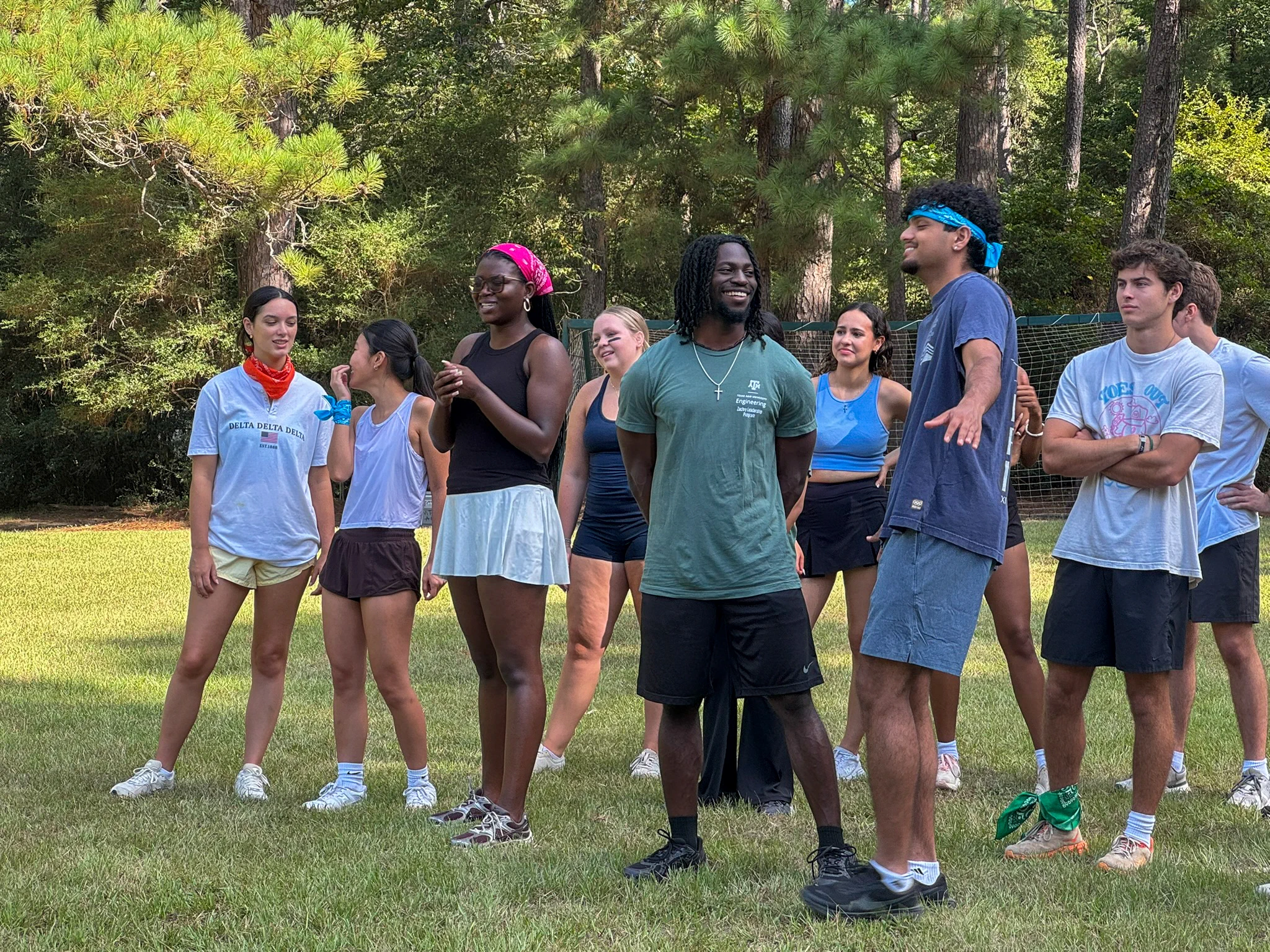 A group of people standing on a grassy field, surrounded by trees. They are casually dressed, wearing athletic clothes and sneakers, and some have bandanas on their heads. They appear to be in a relaxed, social setting.