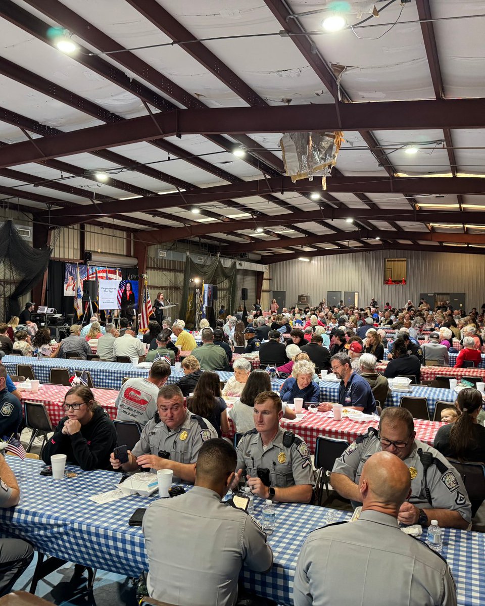 A large indoor community gathering in a metal-roofed hall. Many people sit at long tables covered with red, white, and blue checkered tablecloths, eating and talking. Several uniformed officers sit at a foreground table, while a speaker and display screen are visible near the front. Text appears on the screen and on some signs.