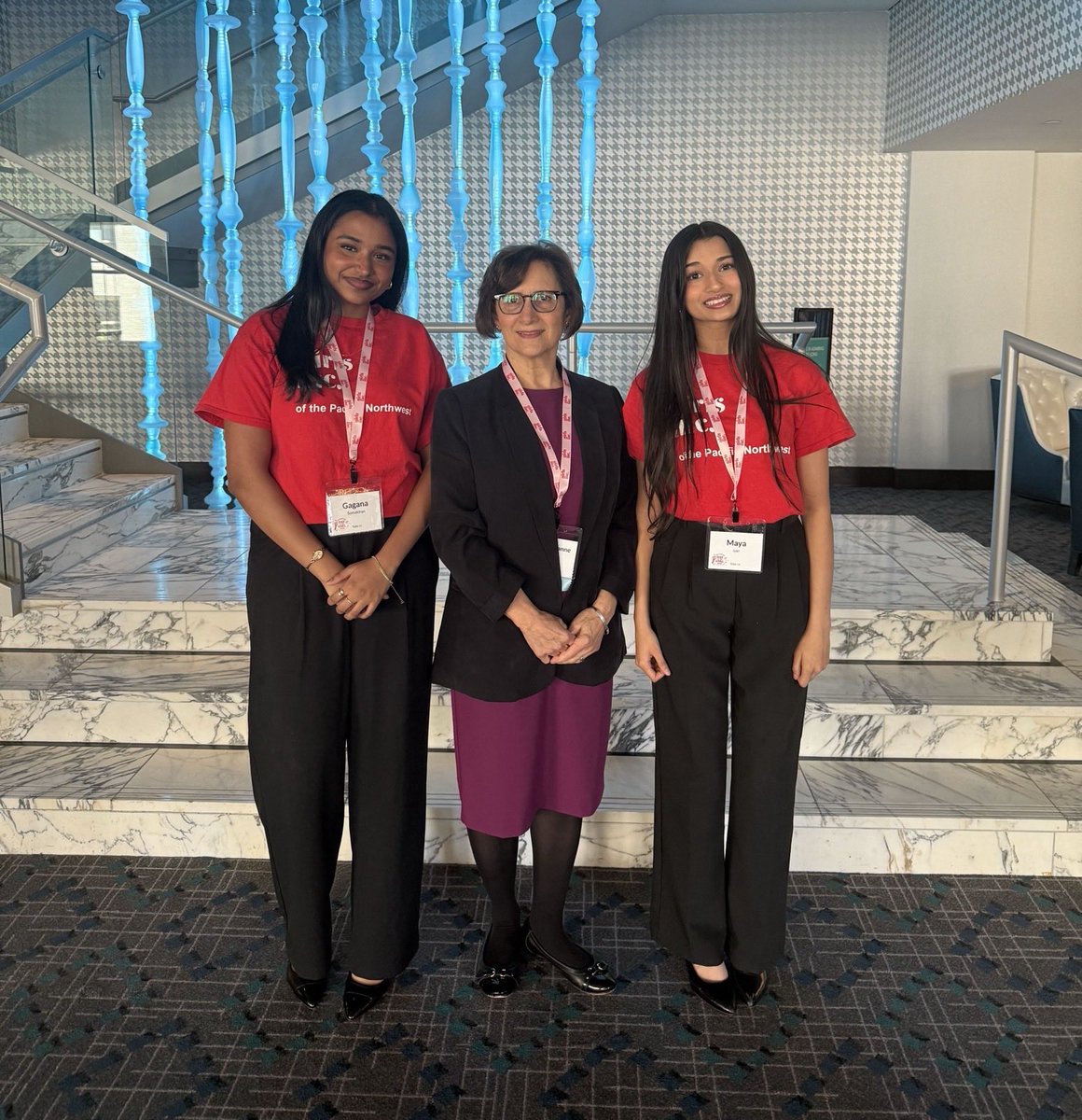 Three women pose together on a marble staircase in an indoor venue. Two wear red shirts and name badges, while the woman in the middle wears a dark jacket and purple skirt. Blue decorative railing posts and patterned walls are behind them. Text appears on their shirts and badges.