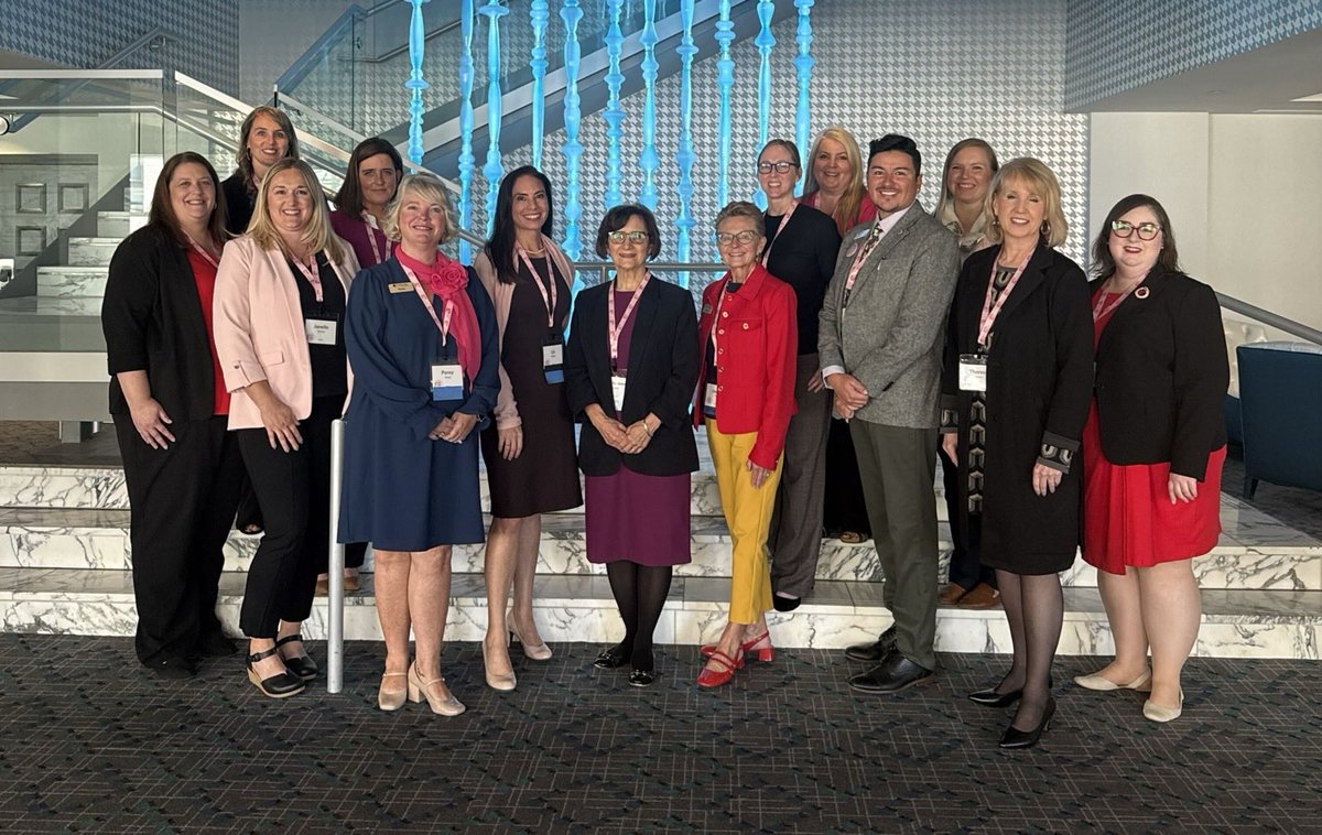 A group of people pose together on marble steps inside a modern building, with a blue decorative installation behind them. Some wear conference-style badges. There is small text on the badges.