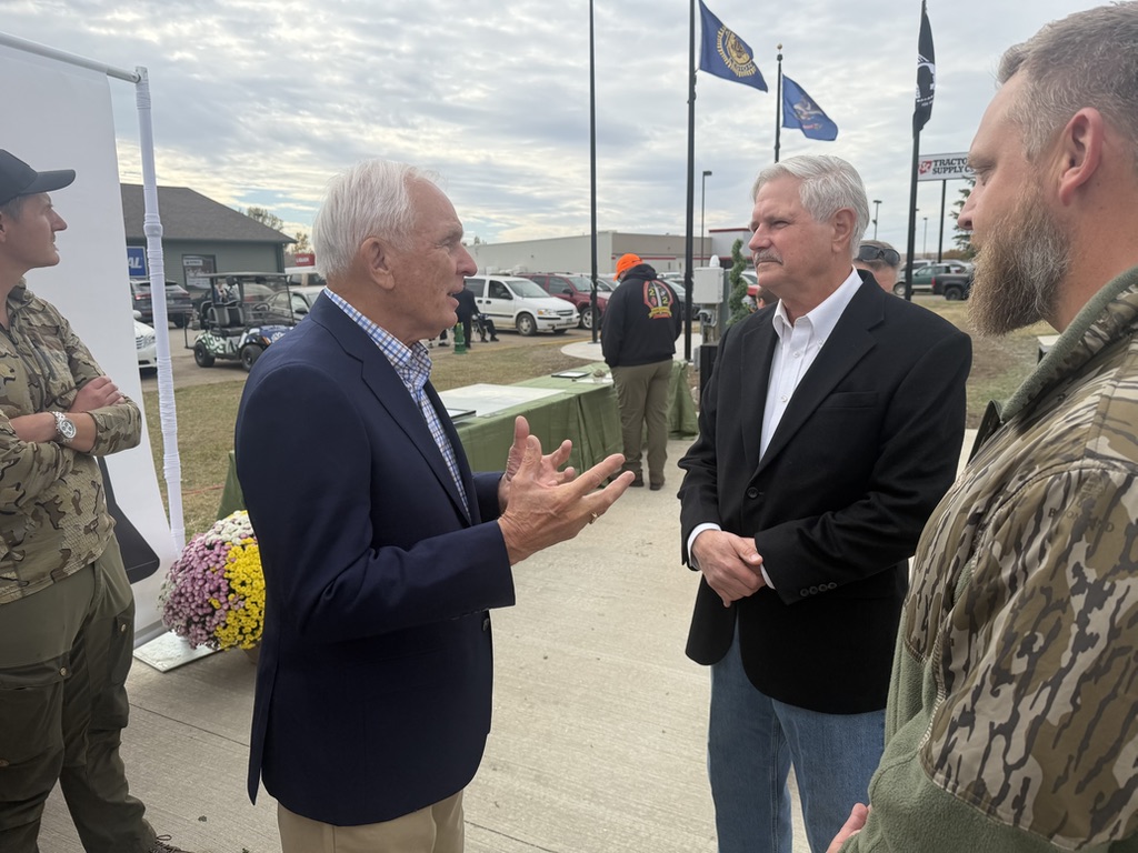 Several men talk outdoors at an event near a table, parked vehicles, and flags. Two men wear camouflage clothing, while another gestures as he speaks to a man in a dark jacket. There is text visible on signs and flags.