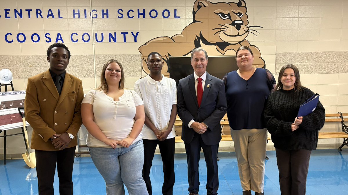A group of six people pose for a photo in a school gym. A large wildcat mascot illustration and school lettering are visible on the wall behind them. There is also a display board at the left with text.