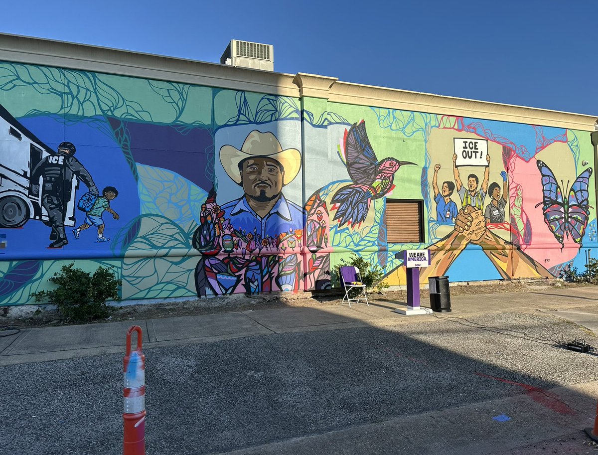 A large, colorful mural covers the side of a building. It depicts a person in a cowboy hat, a hummingbird and butterflies, a scene of a child near an officer, and people raising their hands and joining hands. There are signs with text, plants, a chair, and a trash can in front of the wall.