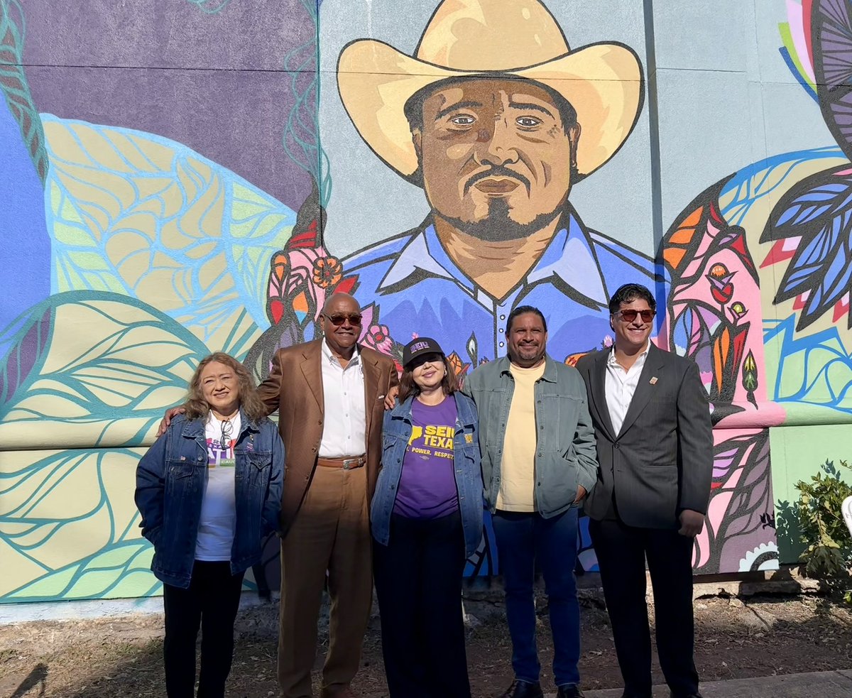 Five people pose together outdoors in front of a large, colorful mural depicting a man in a cowboy hat surrounded by decorative patterns. Some clothing in the image has text.