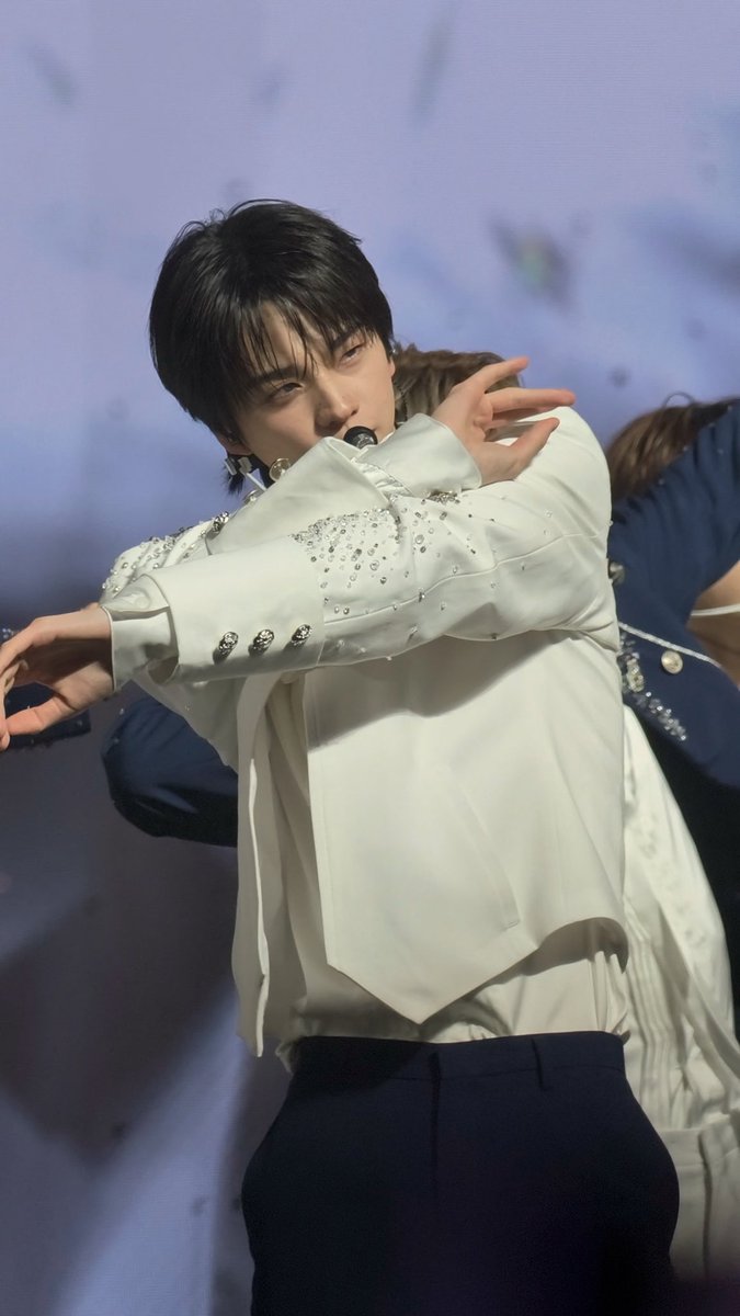 A male performer sings into a microphone onstage, wearing an embellished white jacket and dark trousers, with dancers in navy outfits behind him.