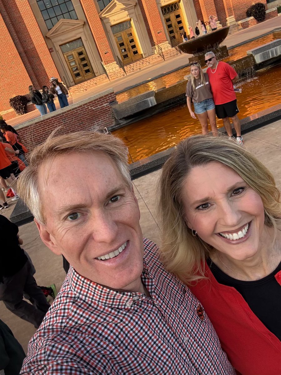 A smiling man and woman take a close-up selfie outdoors beside a reflecting pool and fountain, with a large brick building and other visitors in the background. There is no prominent text.