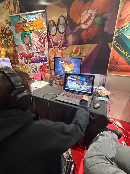 Fotograf铆a a un stand colorido. En el se puede ver a una chica jugar a un videojuego con t铆tulo "Dummies 'n' dochebags" en el pc