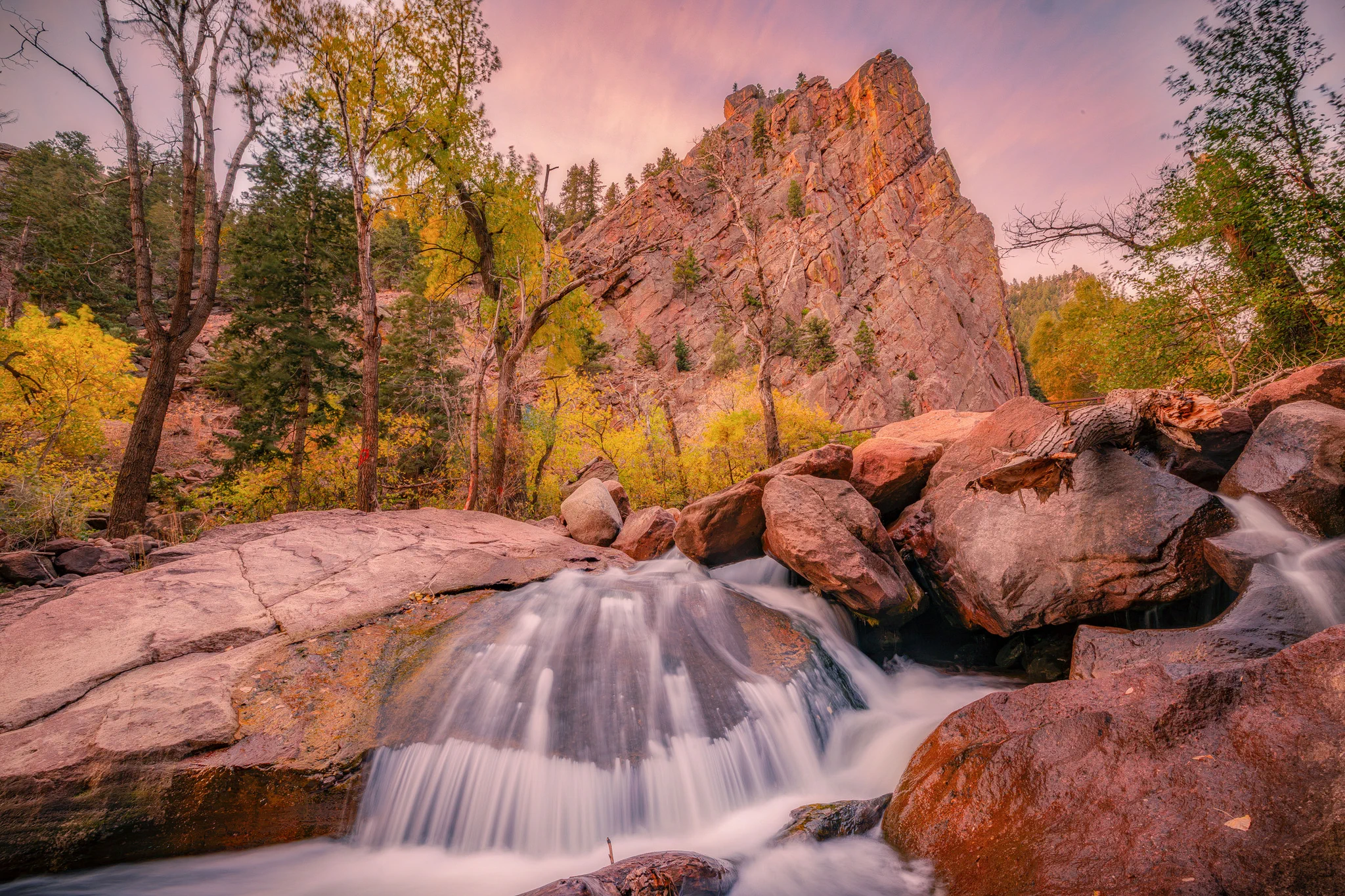 A scenic view of the Boulder Creek at dusk, with smooth flowing water over rocks in the foreground. In the background, the Flatirons rock formations stand tall, surrounded by trees with autumn foliage under a colorful sky.