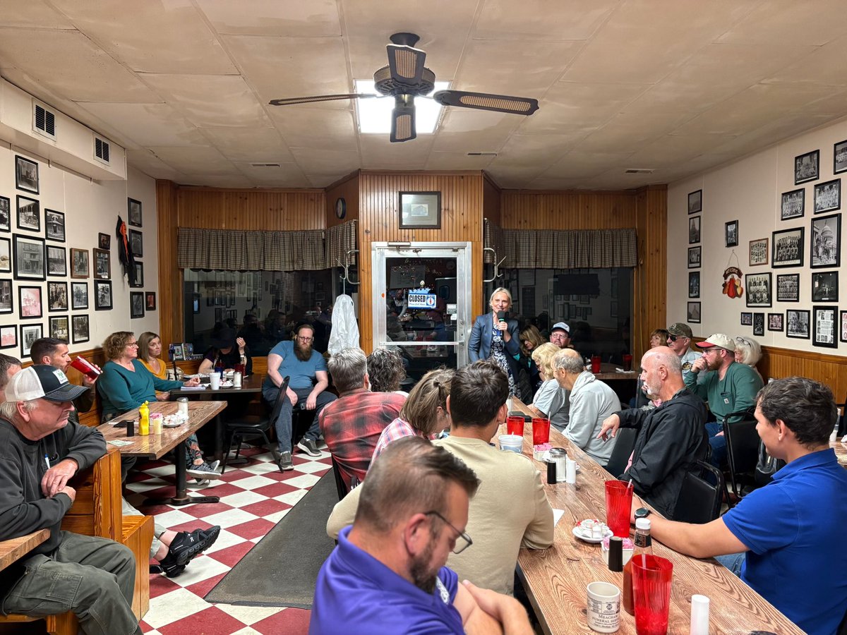 A crowded diner or meeting room where a group of people sit around tables and listen to a standing speaker holding a microphone. The walls are covered with framed photographs, and there is text visible on a sign.