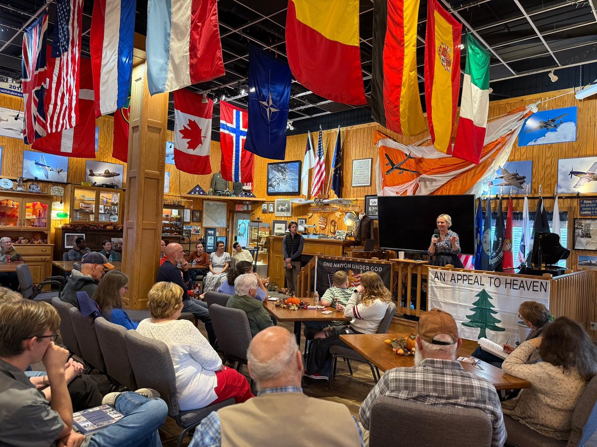 A woman speaks to a seated audience in a room decorated with many national flags, aviation photos, and aircraft memorabilia. A screen and banners are behind her. Text is visible on some banners and signs.