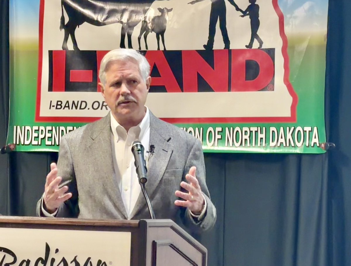 A man in a gray suit speaks behind a lectern with a microphone, gesturing with both hands. A large agricultural organization banner hangs behind him, and there is text on the banner and lectern.