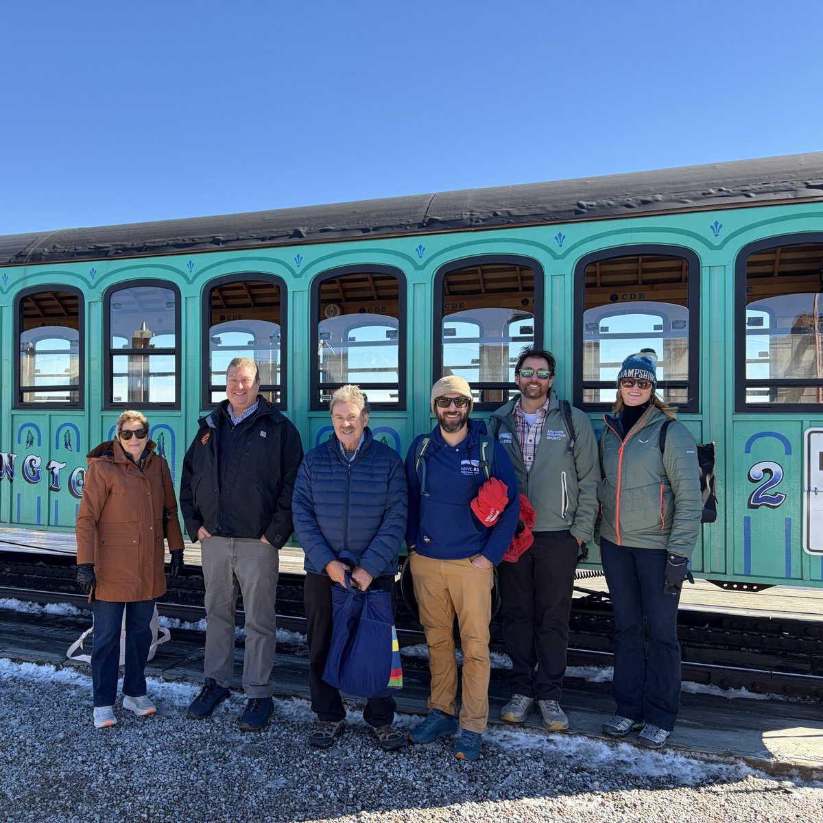 Six people pose for a group photo in front of a turquoise passenger train on a sunny, snowy day. There is lettering and other text on the train and some of the clothing.