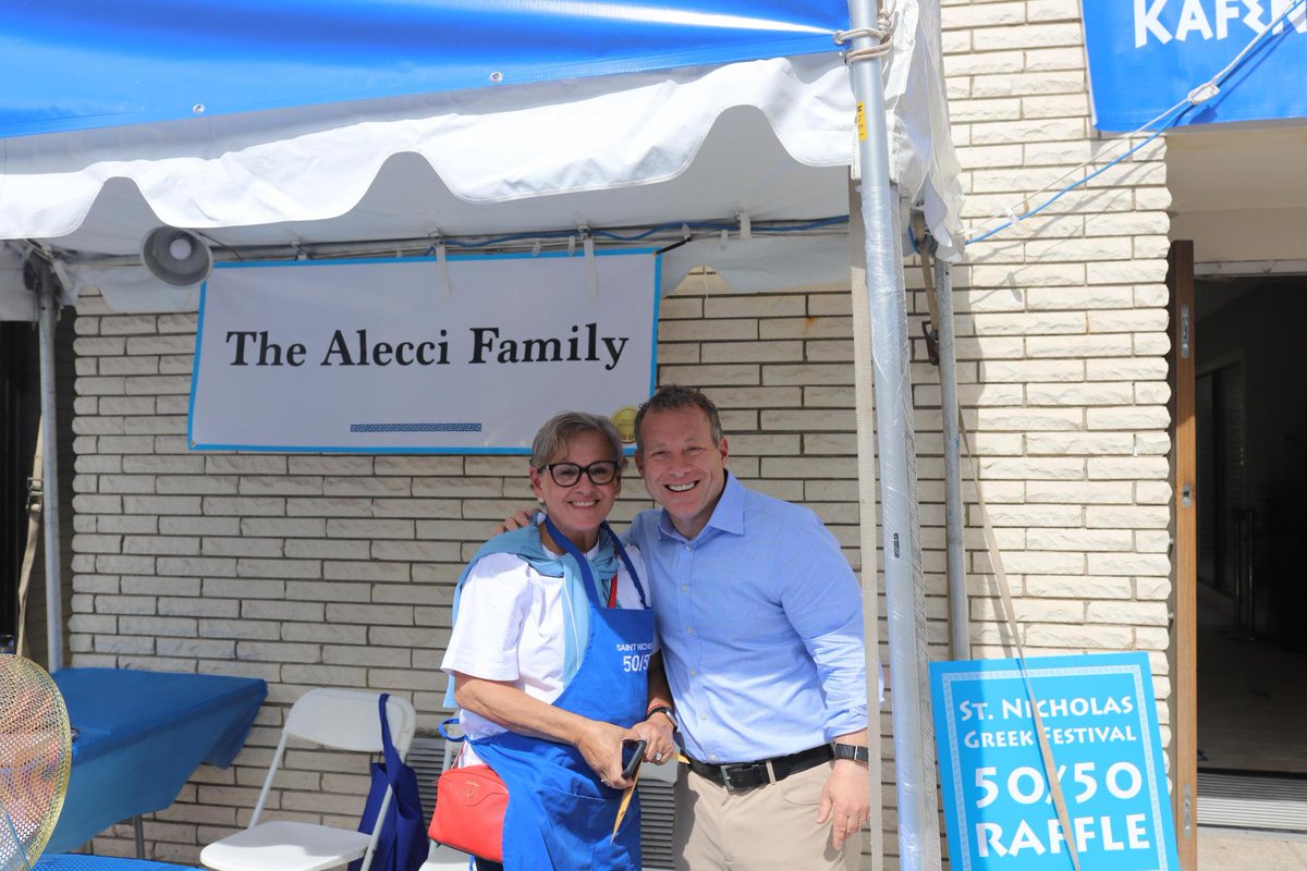 A woman wearing glasses and a blue apron stands beside a smiling man under a canopy at an outdoor festival booth. A brick wall, folding chairs, and signs are behind them. The signs and canopy contain text.