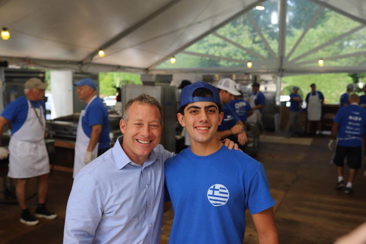 Two smiling men pose together inside a large outdoor event tent, with several people working in the background. One man wears a blue shirt and backward cap, and the other wears a light blue button-up shirt. There is a logo on the blue shirt.