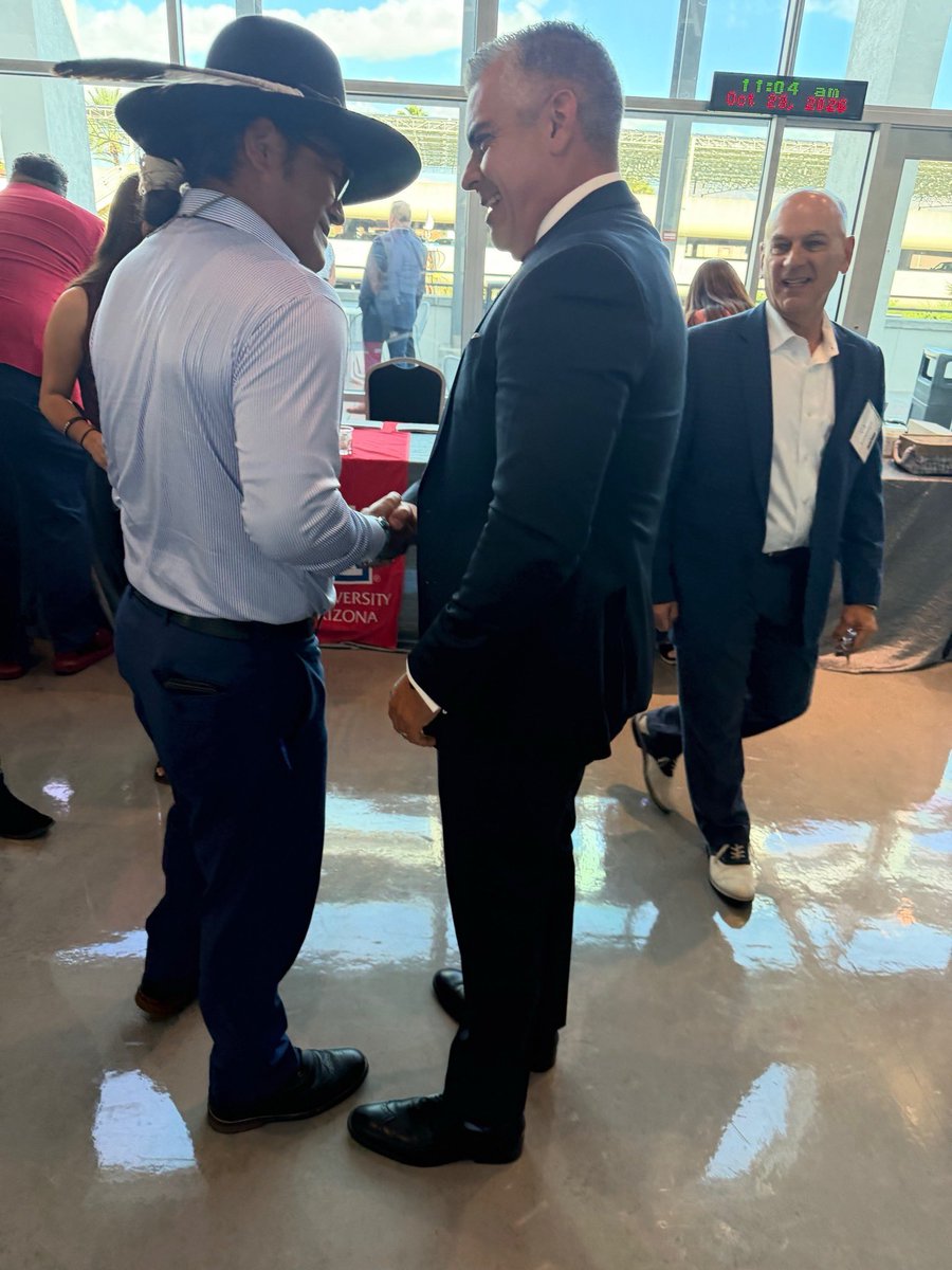 Several people are gathered indoors at an event. Two men in the foreground are talking and shaking hands; one wears a large dark cowboy hat and the other wears a dark suit. Another man stands behind them. Windows, a table, and a digital display with text are visible.