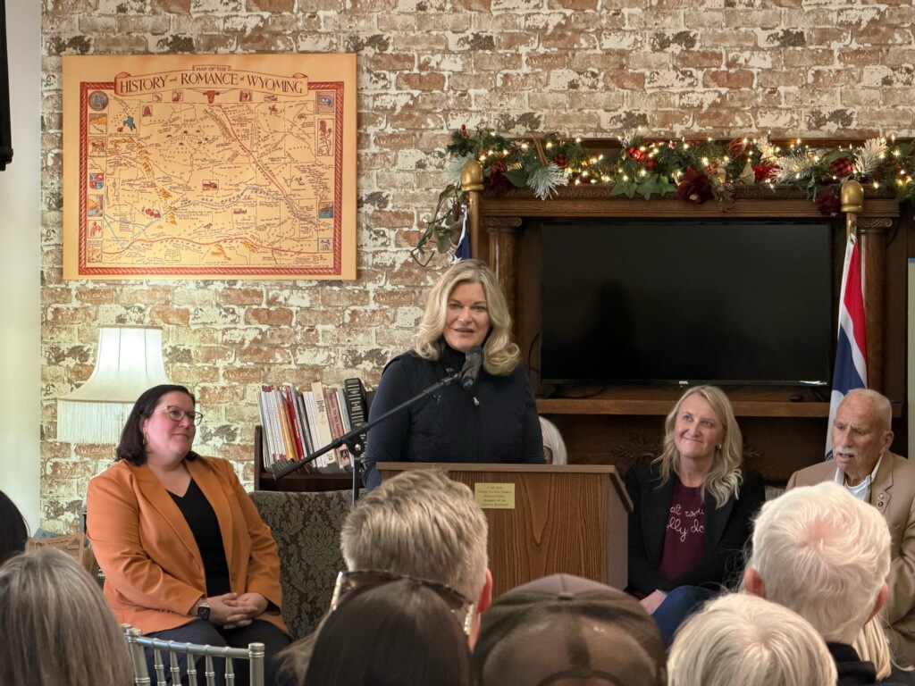 A woman speaks at a wooden lectern to a seated audience in a brick-walled room. Several people sit beside her, with a television, books, framed map, lamp, and holiday garland in the background. Text is visible on the map and a small sign.