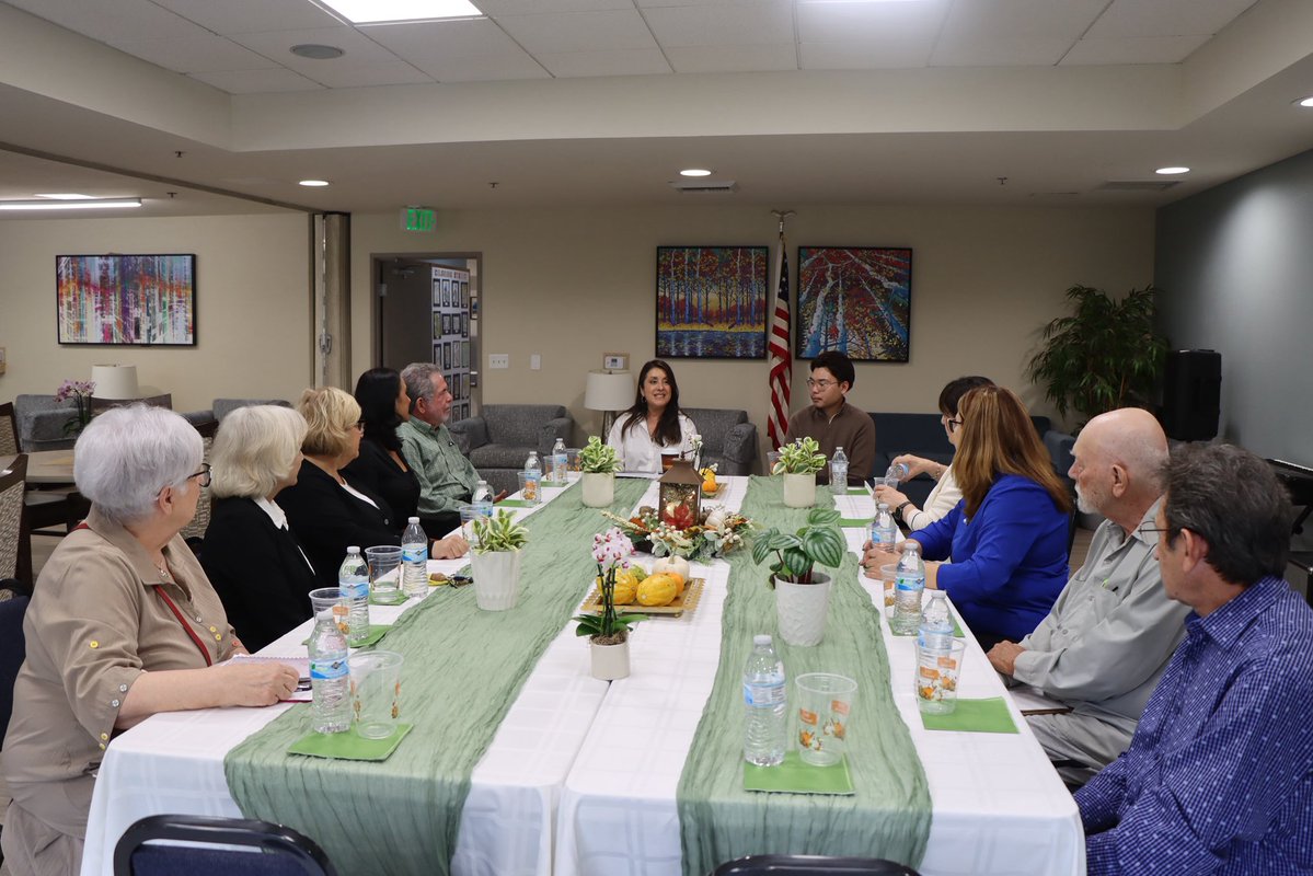 A group of adults sit around a long table in a meeting or community room. The table is set with water bottles, cups, small plants, and food. Wall art and a flag are visible in the background, along with a small amount of text.