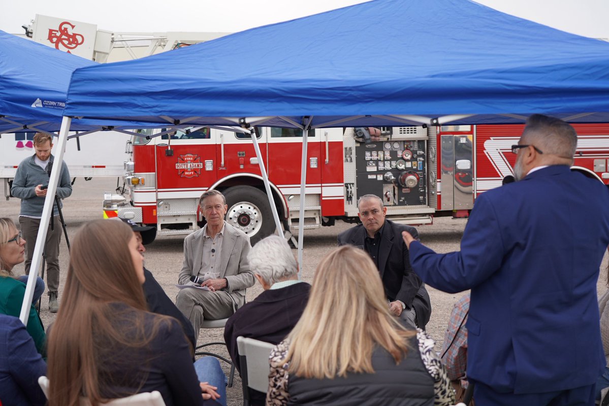 A group of people sit beneath a blue canopy as a man in a blue suit speaks to them. A red fire engine is parked behind the gathering. Text and logos are visible on the fire engine and nearby equipment.