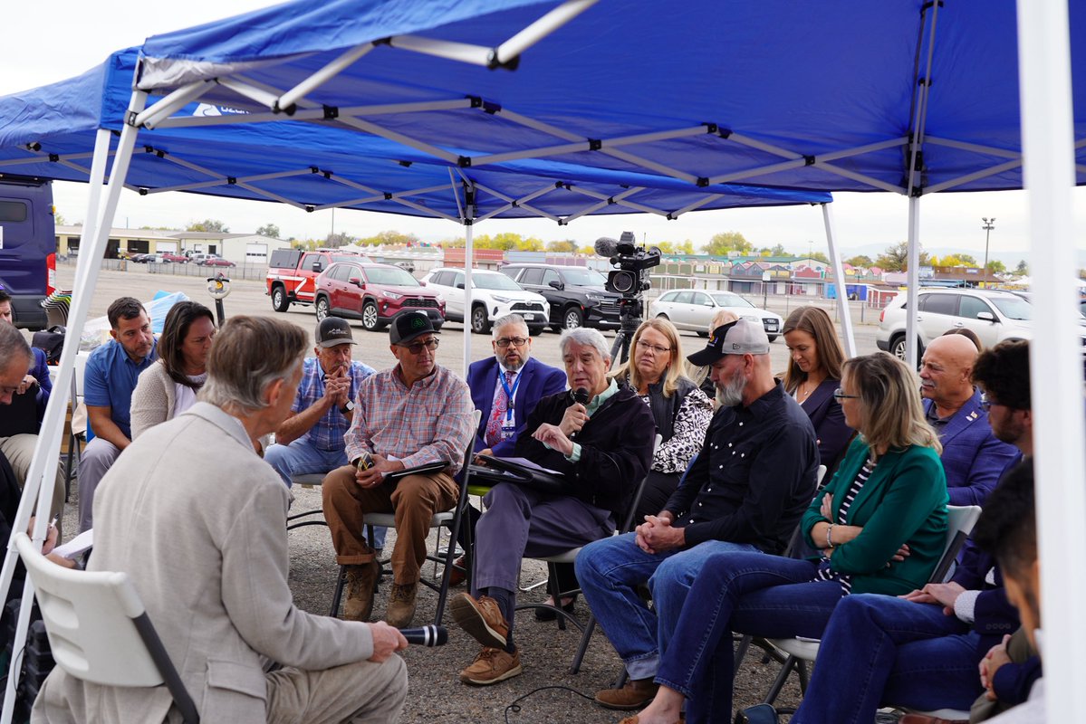 A group of people sits in a circle beneath a blue canopy at an outdoor gathering. One person speaks into a microphone while a camera records the discussion. Vehicles and buildings are visible in the background, and there is some text on the canopy.