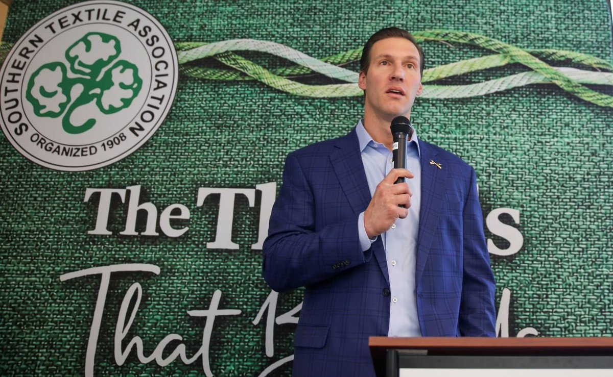 A man in a blue suit speaks into a microphone behind a podium, in front of a green textile-themed backdrop with a circular association emblem and large lettering.