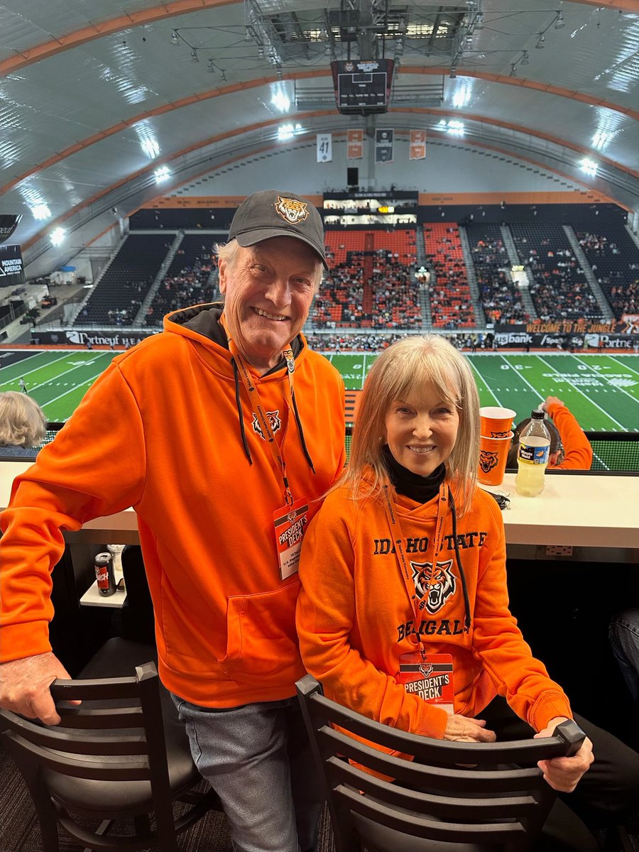 A smiling man and woman pose in orange Idaho State clothing inside a domed football stadium. They are standing by chairs and a counter, with the field, seating, scoreboard, and a few spectators in the background. Text appears on their clothing, badges, and stadium signage.