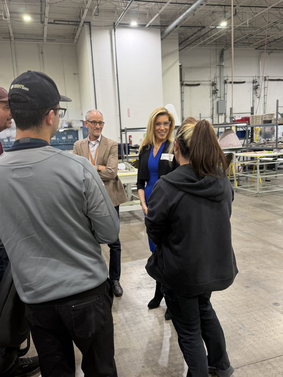 A group of people are talking inside a large industrial warehouse or factory. Worktables, equipment, and exposed ceiling structures are visible in the background. Some clothing and badges contain text.
