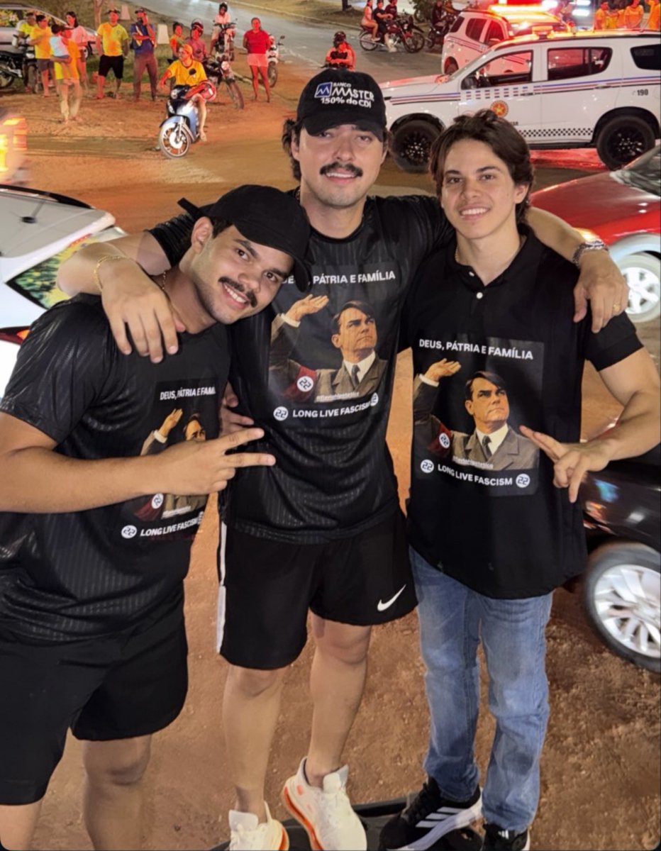 reporterenato's tweet photo. Apoiadores de Flávio Bolsonaro postam foto com apologia ao nazismo na cidade de Grajaú, no Maranhão. Na camiseta, um desenho faz analogia a saudação a Hitler e contém símbolos do nazismo, com a frase "vida longa ao fascismo", em inglês. A imagem foi publicada no Instagram. Viaturas da polícia estavam presentes no ato e aparecem na imagem.