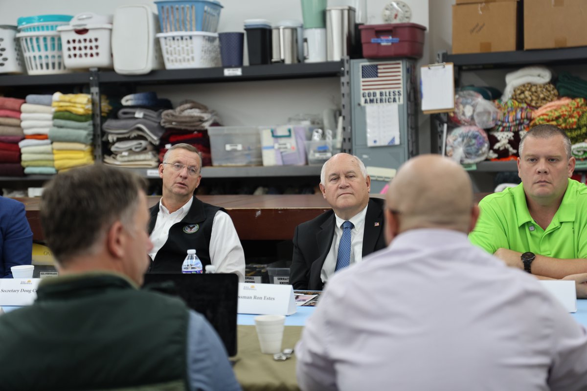 Several men sit around a table in a supply room, appearing to take part in a meeting. Shelves behind them hold folded towels, baskets, containers, and boxes. There is text on a wall poster and on name placards.